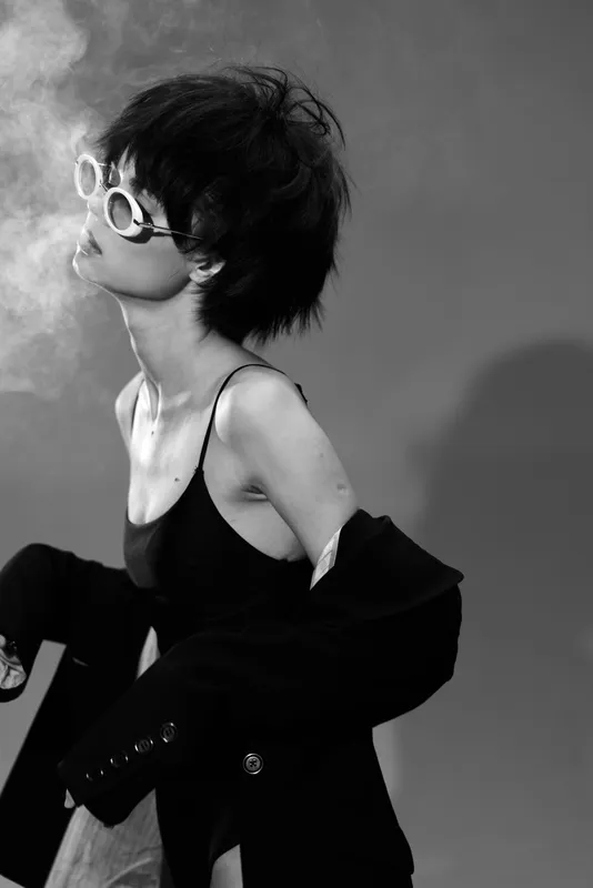 Black and white portrait of a stylish woman wearing sunglasses blowing smoke with edgy fashion and moody lighting