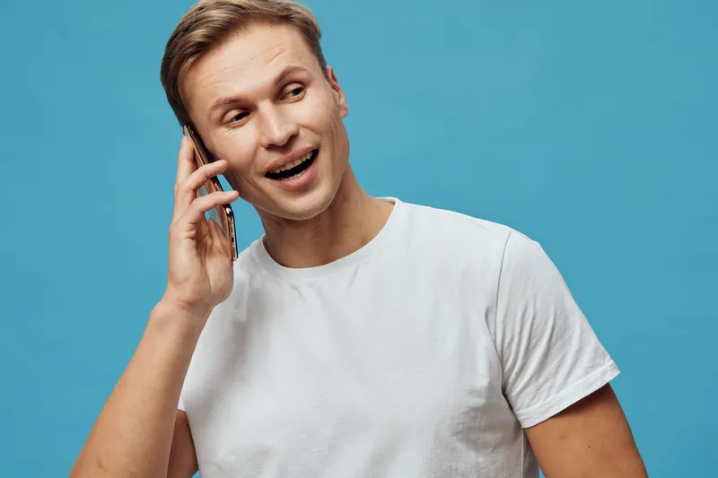 Smiling man talking on mobile phone, casual white t-shirt, cheerful expression, young adult, short hair, isolated on blue background, lifestyle concept, communication, technology, human emotion.