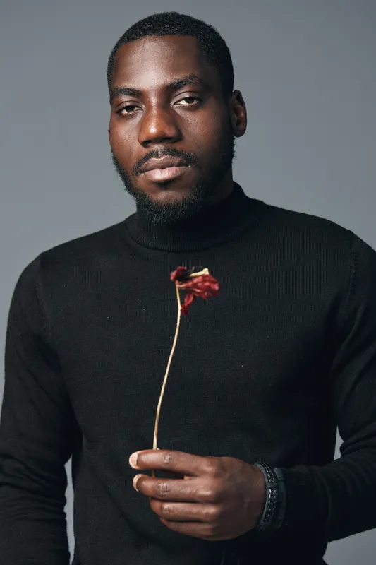 African man in black turtleneck holding dried flower in creative conceptual visual style studio portrait with serious expression and minimalistic gray background.