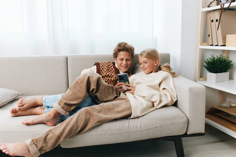 Two smiling children lounging on a light sofa, sharing a smartphone for playful entertainment and relaxed togetherness in a bright, cozy living room.