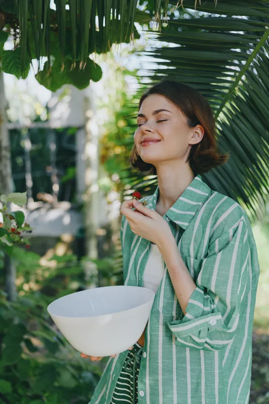 Cheerful woman outdoors in a lush garden, wearing a green striped shirt, holding a bowl, and smiling with calm satisfaction.