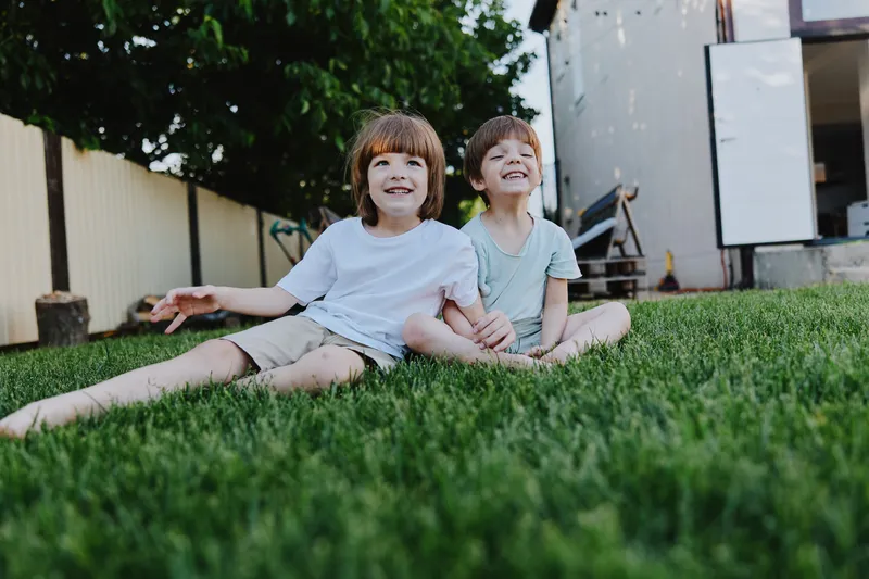 Happy boys sitting on green grass, enjoying a sunny day with cheerful smiles, reflecting friendship and joy of childhood in a serene backyard setting.