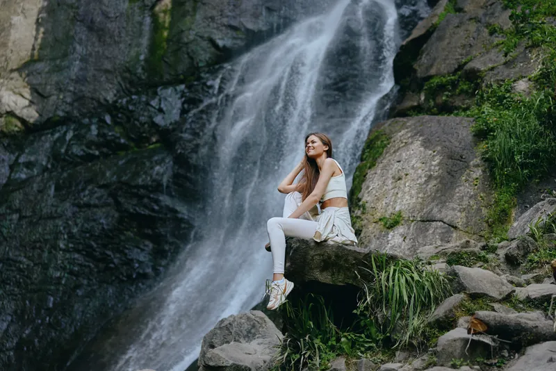 Young woman sitting on rock near waterfall, wearing casual white outfit, relaxing outdoors in lush green nature during daytime, peaceful and serene atmosphere.