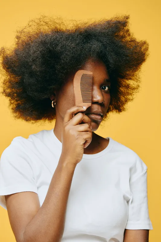 African woman with natural hair holding a comb, showing confidence against a vibrant yellow background, embracing natural beauty and self care