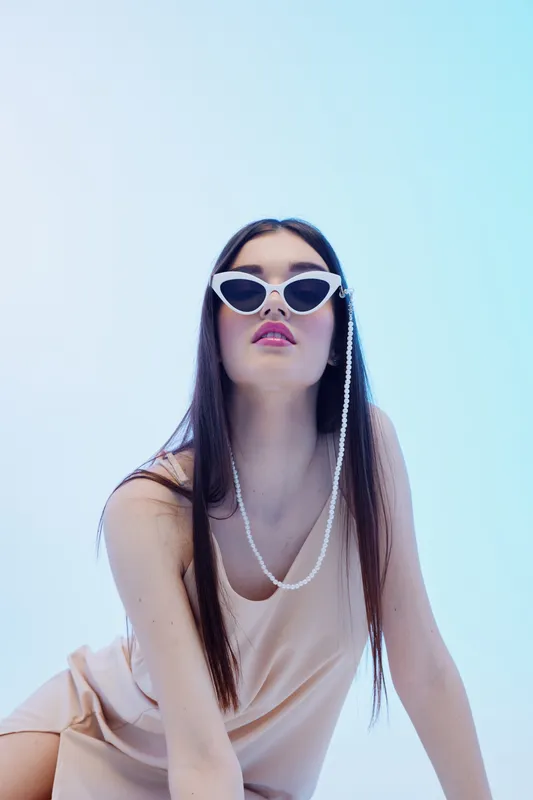 Young woman with long dark hair wearing stylish white sunglasses and a beige dress poses confidently against a light blue background. Concept of beauty fashion and elegance.