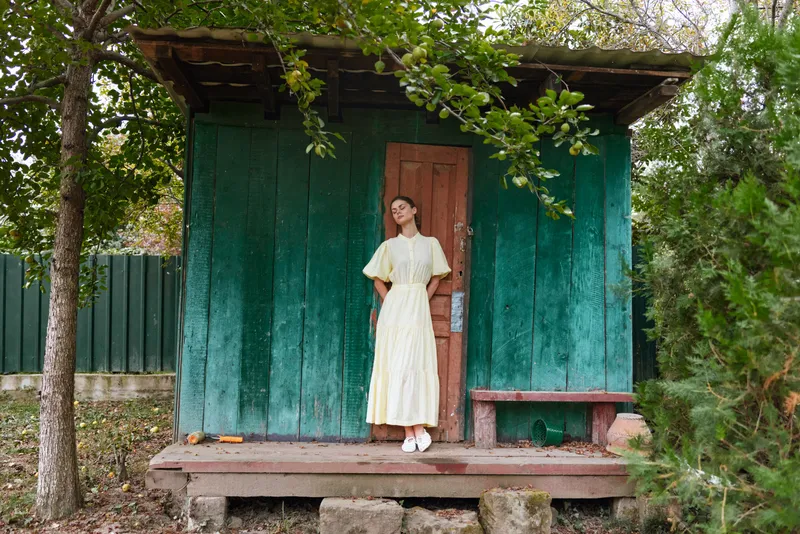 Woman cottage vintage portrait in light dress standing at rustic turquoise doorway, outdoor scene with authenticity, inclusivity, rural cabin entrance and foliage background