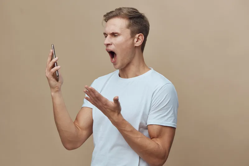 frustrated man holding smartphone yelling in white t-shirt isolated on beige background cybersecurity concept