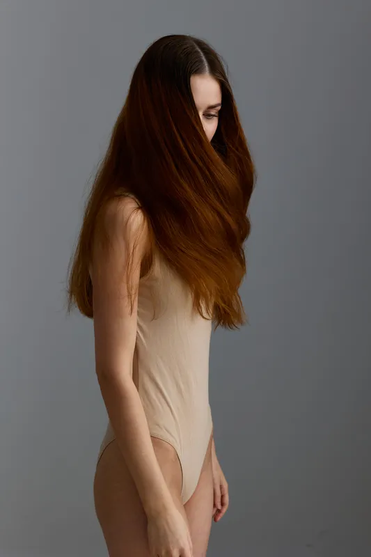 young woman with long brown hair wearing beige bodysuit stands against neutral gray background, natural light enhancing smooth skin and delicate features, minimalistic.