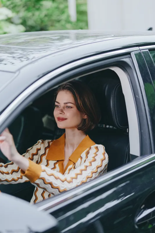 Portrait of a woman driving a car, hands on the wheel, calm expression, wearing a striped knit sweater, sunlight on the dashboard.
