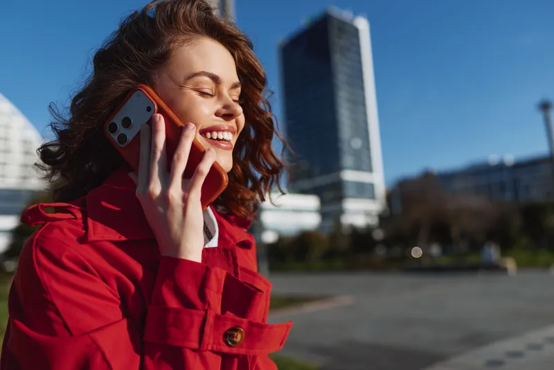 Young woman with curly hair wearing a red coat talks on a phone in a sunny urban plaza with tall buildings in the background, radiating confidence and casual style.