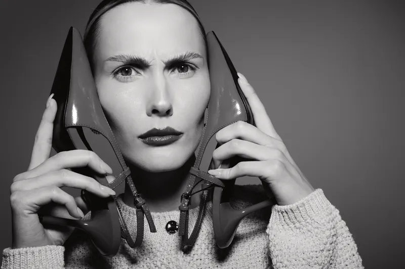 Confused white woman with pale skin holds two high heel shoes near her face, wearing knitted sweater in studio. Concept of fashion dilemma, footwear choice, and style confusion.