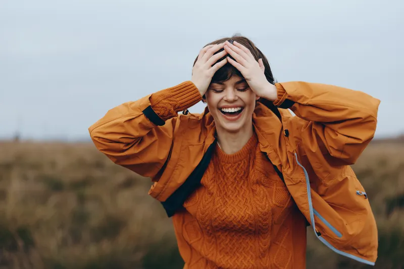 Woman outdoors laughing with both hands on head, orange jacket and sweater, autumn field background, sunlit breezy moment, relaxed lifestyle, colors, soft focus, portrait, shallow depth