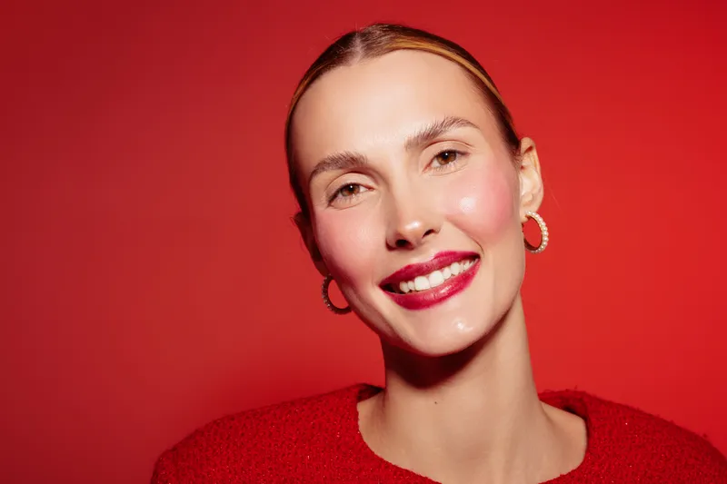 Smiling woman with red lipstick and hoop earrings poses against red background. Close-up portrait with natural skin and bright makeup. Concept of beauty and confidence.