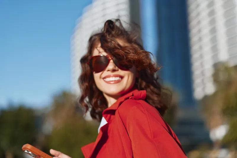 Smiling woman with curly brown hair wearing a red jacket and sunglasses, posing outdoors in an urban setting with modern buildings behind, bathed in warm light.