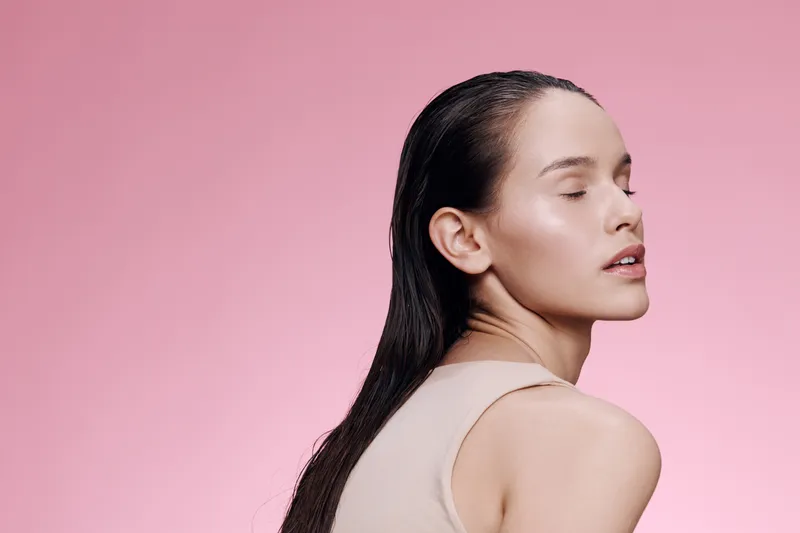 Young woman with long hair on a pink background, expressing calmness and confidence, wearing a simple sleeveless top, ideal for beauty and wellness concepts