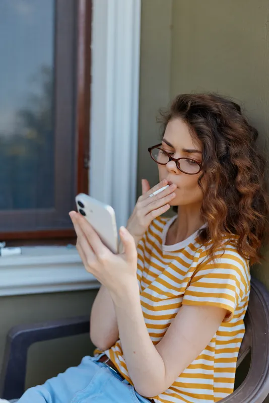 A woman with glasses in a yellow striped t shirt sits on a balcony using a smartphone, lighting a cigarette. A candid home lifestyle moment highlighting personal time and casual fashion.