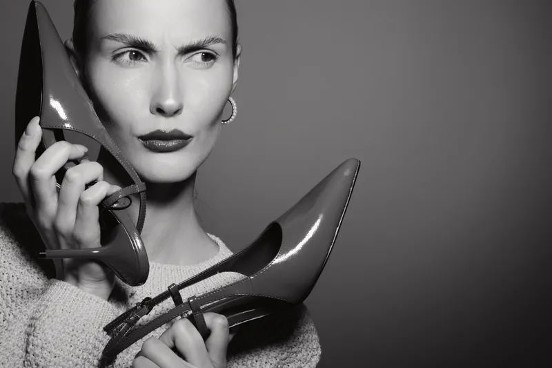 Confused Caucasian woman with pale skin holding high heel shoes near face in studio. Concept for fashion choice and shopping dilemma.