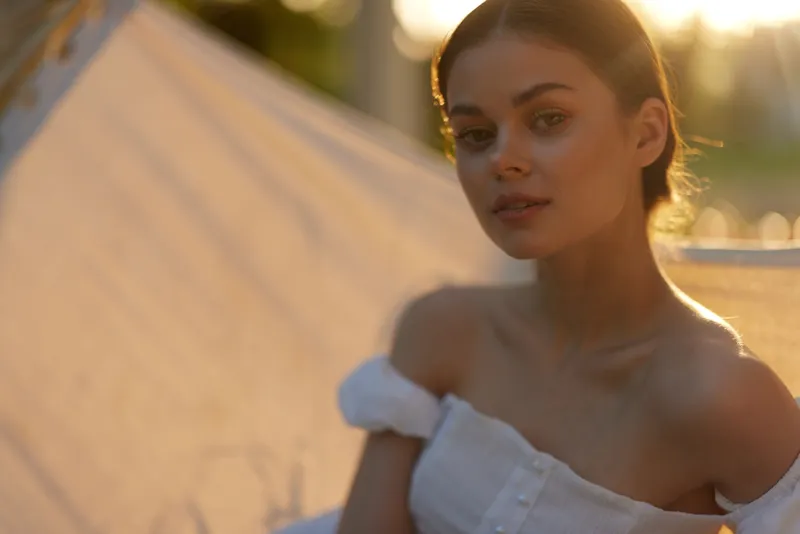 Young woman in a white off shoulder dress with a serene expression, photographed during golden hour, capturing a warm and inviting atmosphere