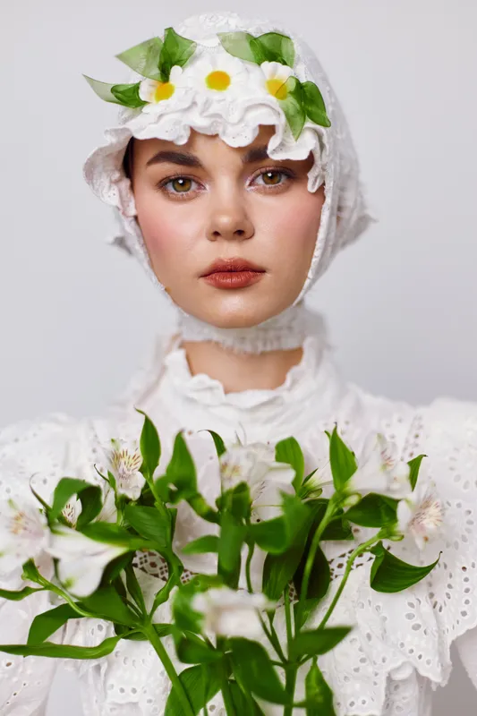 Ethereal young woman in a white vintage dress holding flowers, showcasing natural beauty and serenity in a soft, floral themed setting