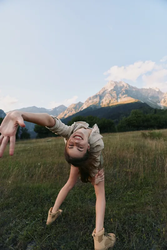 Vibrant woman lifestyle moment outdoors in a sunny meadow with mountains in the background, playful movement and candid joy, natural colors and fresh air.