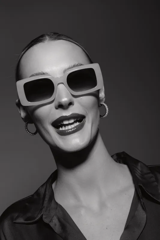 Smiling white woman with light skin wearing oversized sunglasses and hoop earrings against dark background.