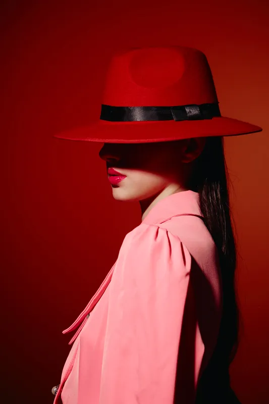 Stylish woman in red wide-brim hat and pink coat poses against red background. Side profile shows bold red lips and sleek dark hair. Concept of beauty, fashion, and creative style.