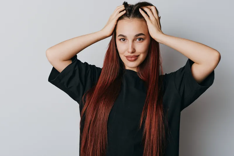 Young woman with long red hair and black shirt posing with hands on head smiling against white background. Portrait of confident female with natural makeup and casual style.