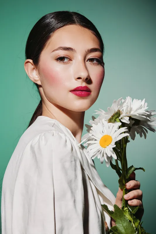 Young woman with long dark hair holds white daisies close to her face. Her bright red lips contrast with soft makeup and simple white shirt on green background. Beauty creative concept.