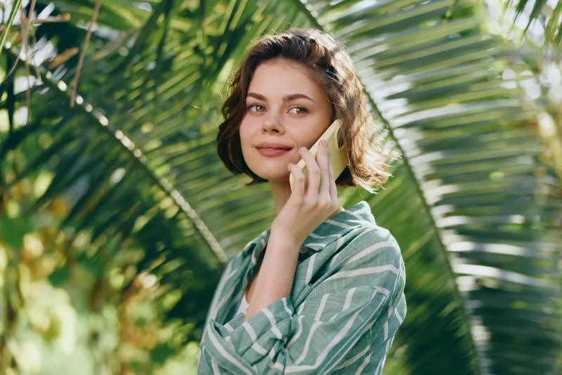 Portrait of a young woman outdoors, talking on a smartphone amid lush tropical greenery.
