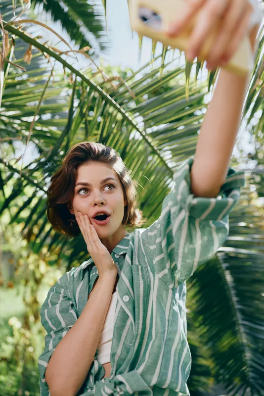 Young woman takes a carefree selfie outdoors among tropical palm leaves, her mouth open in astonishment and a delighted expression.