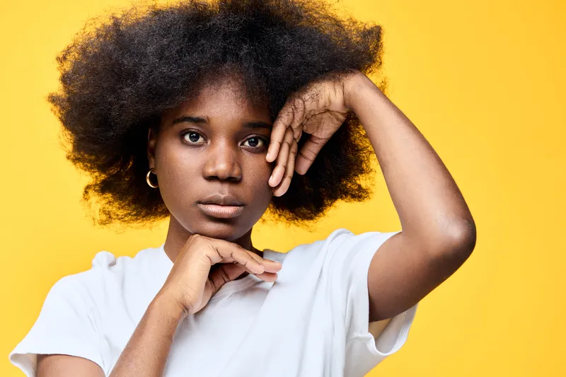 A close up portrait of a young African woman with natural curly hair, wearing a plain white shirt against a vibrant yellow background, conveying a sense of confidence and elegance