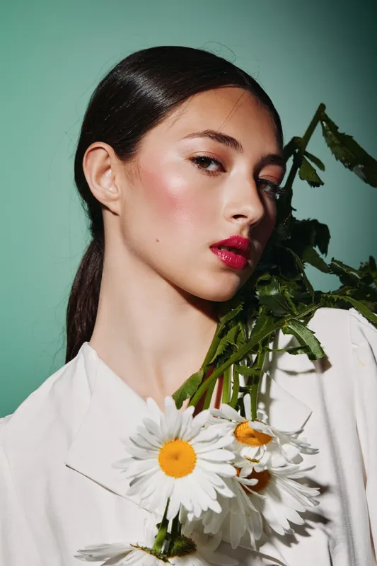 Young woman with slick dark hair holds chamomile flowers on bright green background. She wears glossy lipstick and soft makeup, creating a modern beauty and fashion concept.