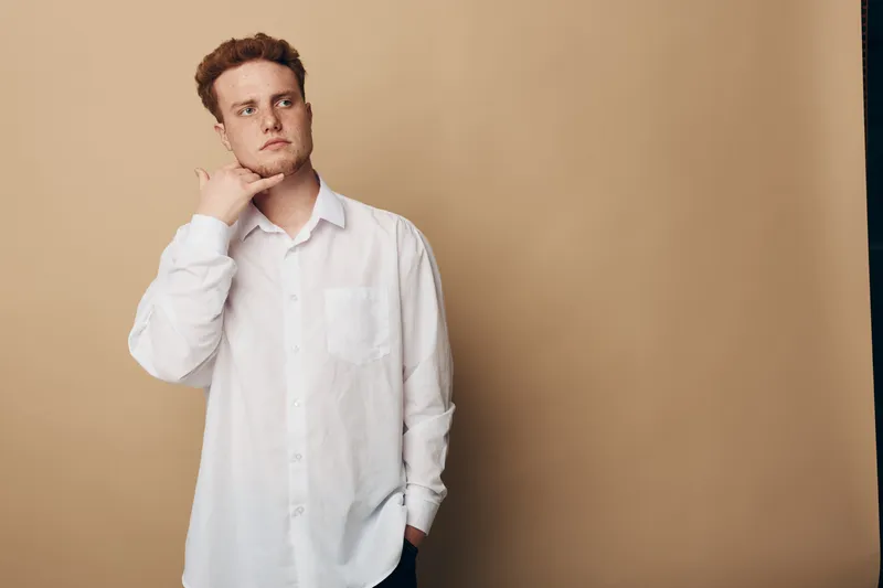 Young man in white shirt makes call me gesture with hand near face, looking away on beige background. Concept for communication, phone call, gesture, and casual style.