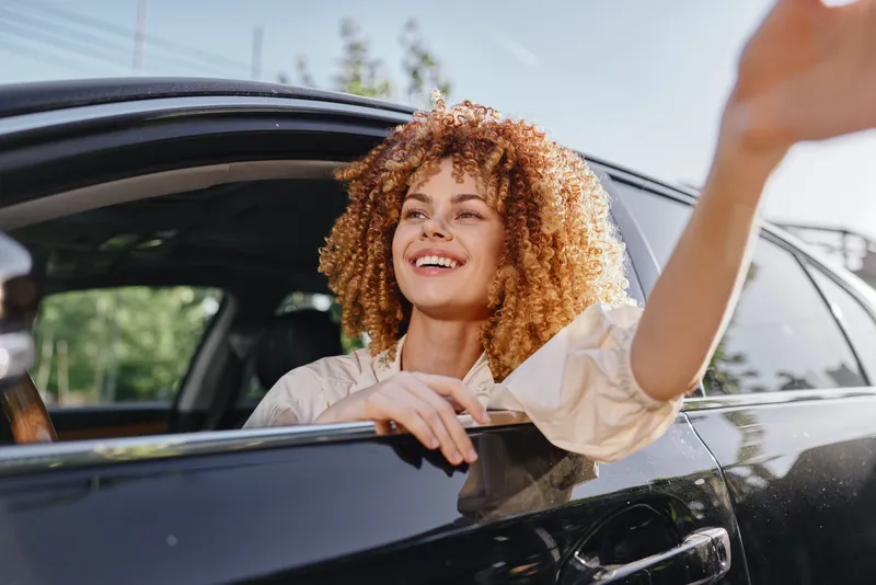 smiling woman with curly hair waving hand from black car window on a sunny day, casual style, cheerful mood, outdoors, modern urban setting