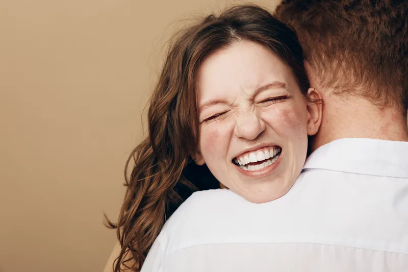 Happy woman with closed eyes and wide smile embraces man wearing white shirt. Warm beige background creates cozy atmosphere. Concept of love, affection, and joyful relationship.