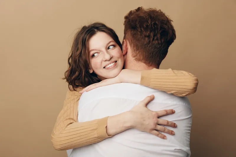 Smiling woman with brown hair hugs man wearing white shirt on beige background. Intimate moment of affection and connection between couple. Concept of love, relationship, and happiness.