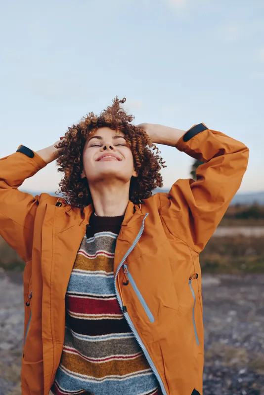 Woman outdoors, smiling with eyes closed, curly hair in wind, orange jacket and striped sweater, hands behind head, warm sunlight, relaxed lifestyle, open road landscape, natural colors