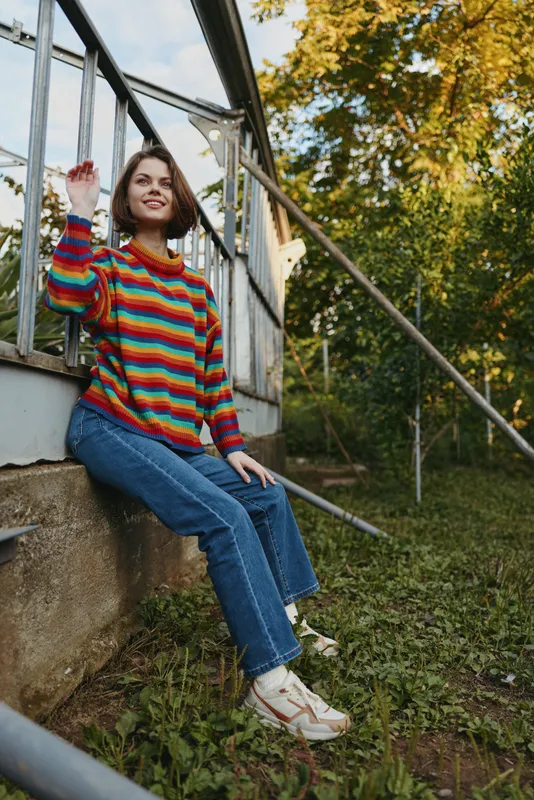 Woman in colorful striped sweater sitting on a greenhouse ledge, smiling and relaxed in jeans and sneakers outdoors among autumn trees and grass, casual garden portrait.
