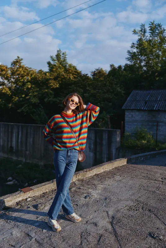 Young woman and woman in a casual outfit — striped sweater, jeans and sunglasses — smiling with a crossbody bag while leaning outdoors on a rural roadside fence for relaxed travel lifestyle.