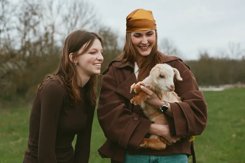 Two women outdoors with a kid goat, smiling and relaxed, sharing a warm lifestyle moment as they hold the young animal in a green field, wearing casual jackets, under an overcast sky, with a friendly