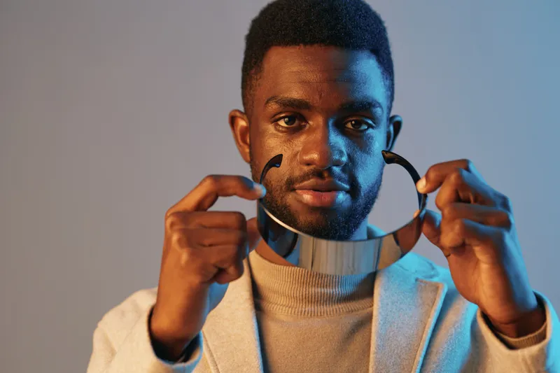 A stylish male model of African descent holding a modern reflective collar, wearing a light sweater and blazer against a blue background, showcasing a confident and creative vibe