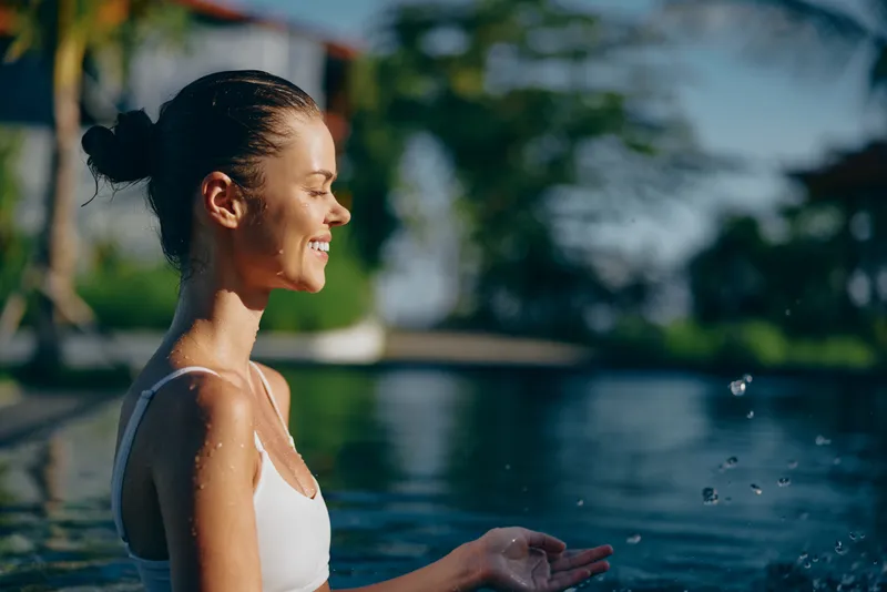 Smiling woman in a white tank top stands beside a sunlit pool, water droplets glistening in the air. Concept of wellness, relaxation, and outdoor lifestyle with bright, natural lighting.