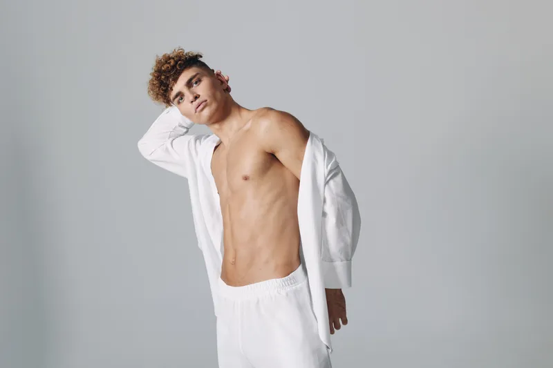 Young man with curly hair posing shirtless in white pants and open jacket against plain background. Fashion model with toned body and casual style in studio lighting.