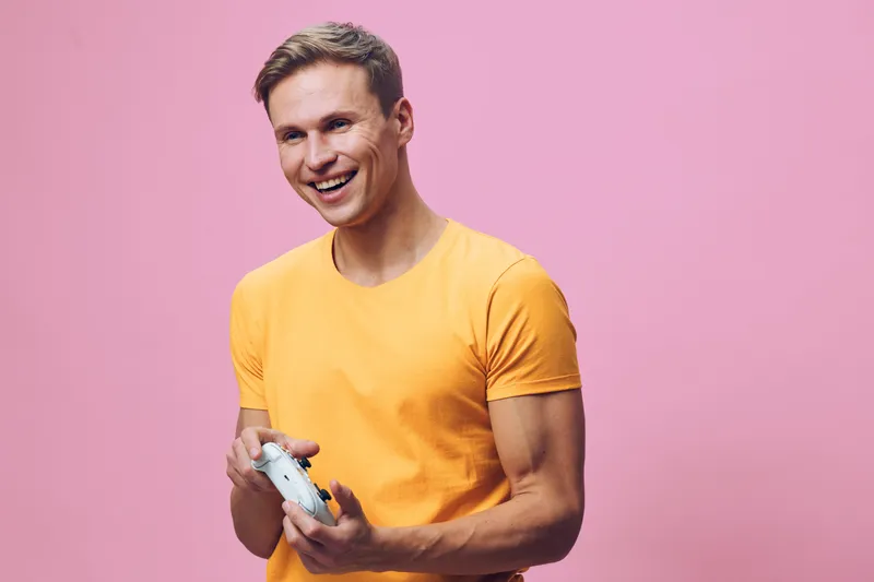 Cheerful young man in bright yellow t-shirt holding video game controller smiling, isolated on pink background. Casual lifestyle entertainment fun enjoyment leisure concept.