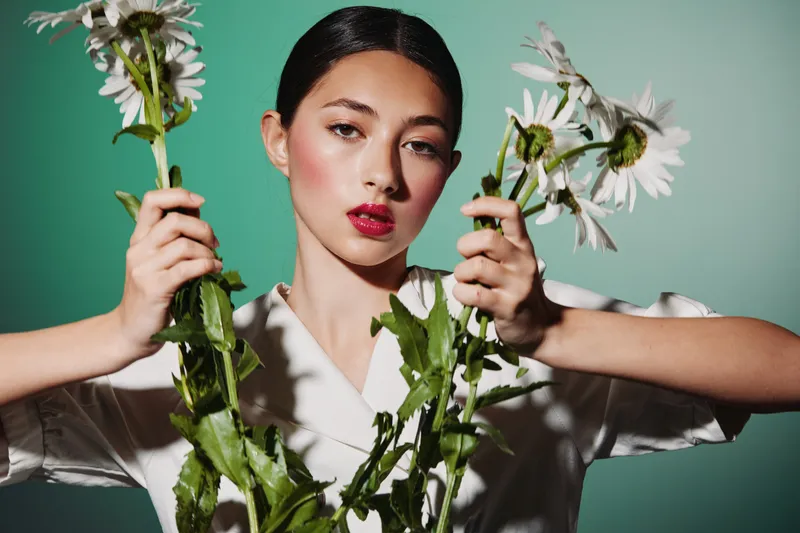 Young woman with slick black hair wears chic white top holding large garden flowers against a vintage teal studio background. Concept of beauty, creative, fashion photography.