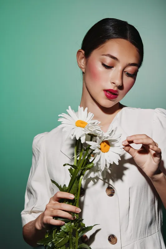 Young woman in a white blouse holding white daisies, looking down with red lipstick and glowing skin. Studio fashion portrait on green background. Concept of beauty creative fashion.