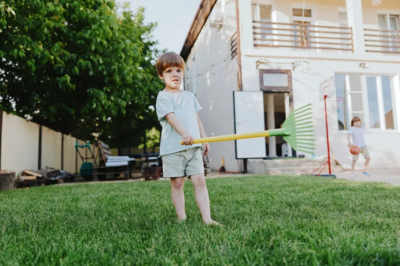 Playful boy holding a rake enjoys a sunny day outdoors, surrounded by green grass and a bright atmosphere, capturing the essence of childhood and fun.