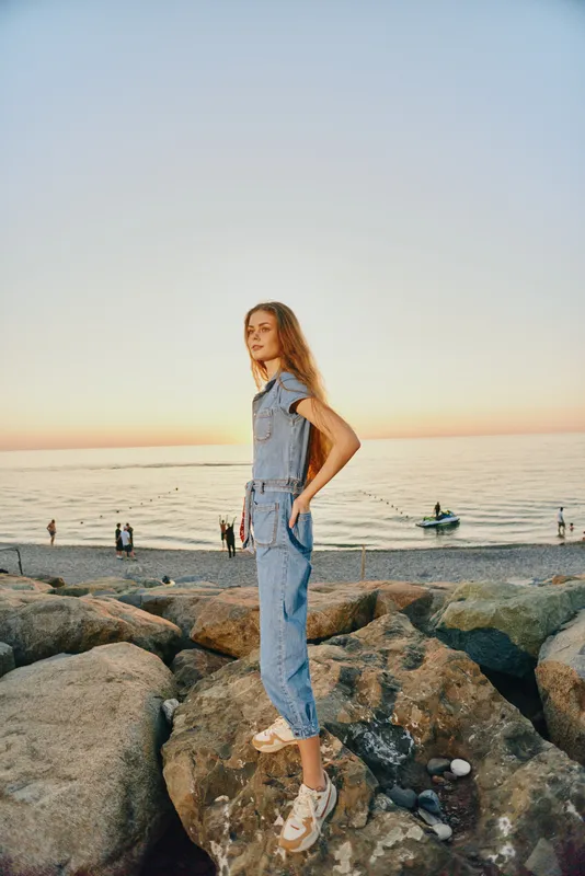 Lifestyle woman in modern boho-western denim street style standing on rocks at sunset by the sea with film color effect and relaxed natural pose.