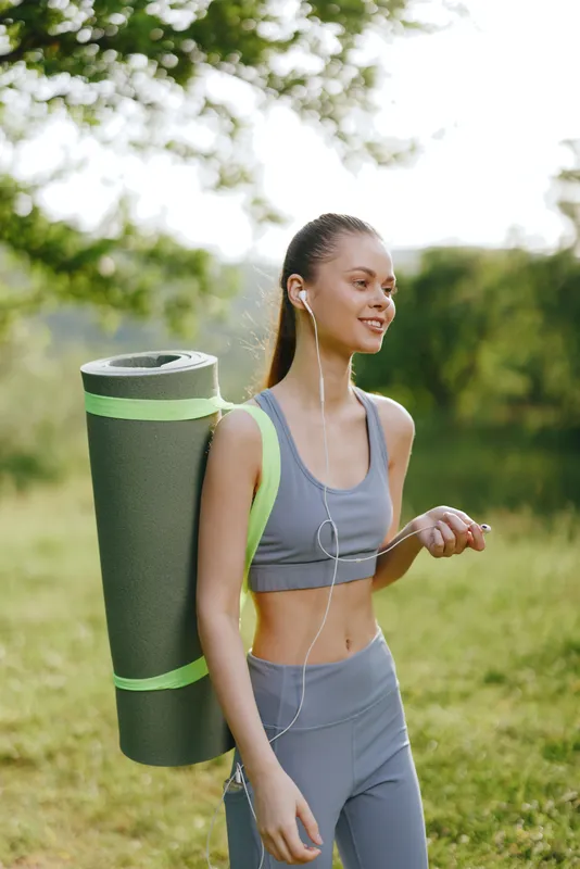 Young woman carrying a yoga mat, dressed in activewear, smiling joyfully in a lush green park, embodying fitness, health, and wellness concepts