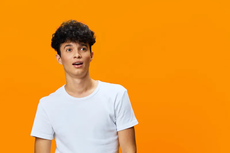 Young man with curly hair wearing white t-shirt looking surprised against bright orange background expressing astonishment and curiosity.